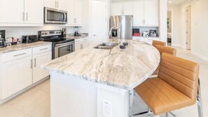 A modern kitchen with white cabinetry, a central island with seating, stainless steel appliances, and a marble countertop. - Amazing 4/3 Home at Reunion!! With Private Pool, Near Disney and Universal!! (Kissimmee)