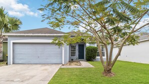A single-story house with a white garage door, a front door, and a small front yard with a tree and grass. - 5/4 Pool Home Located in Sunset Ridge Community! (Davenport)