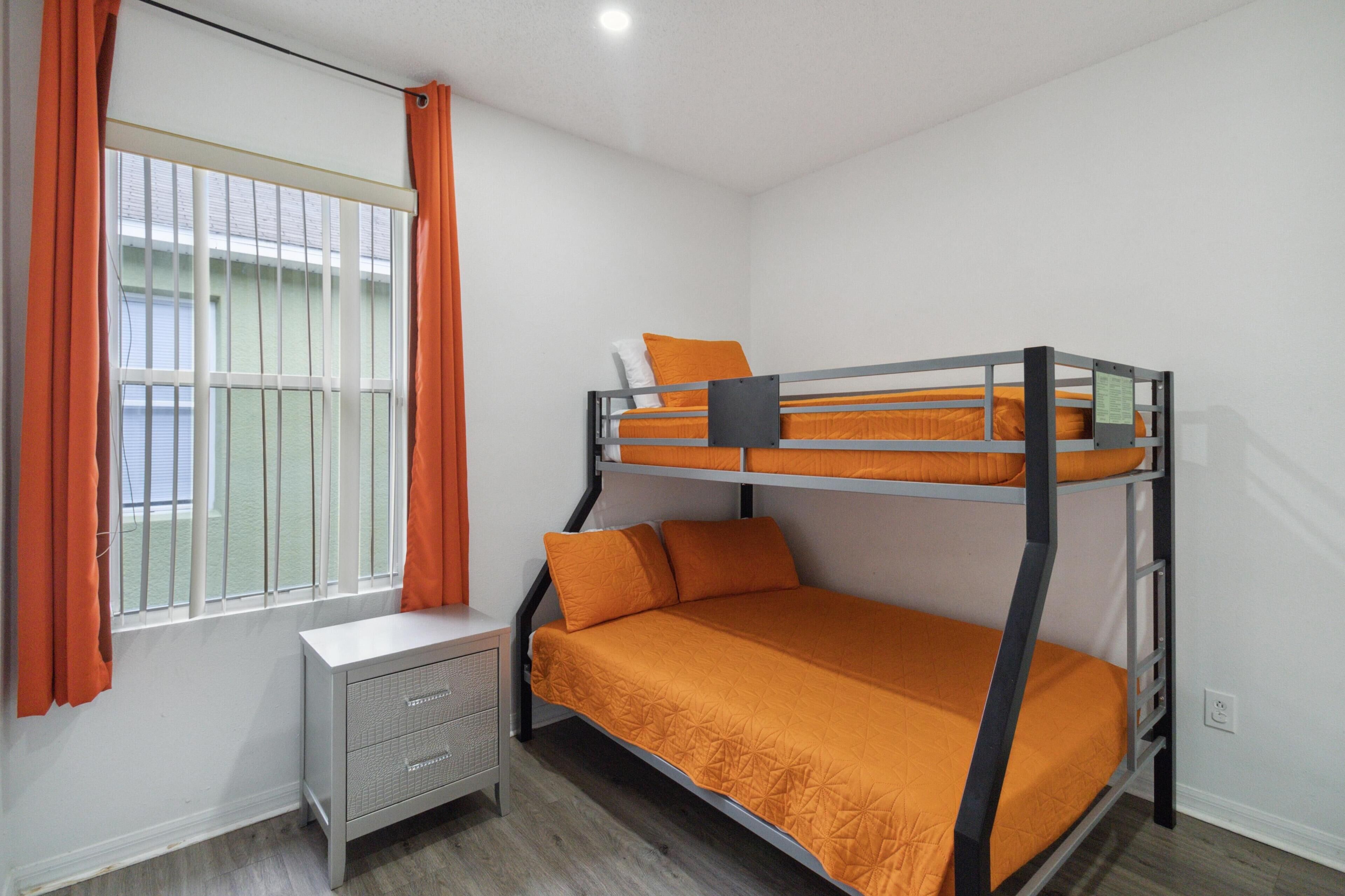 A bunk bed with orange bedding, a small bedside table, and a window with orange curtains.