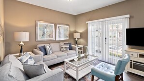 A modern living room with a sofa, coffee table, and TV. There are two framed artworks on the wall and a window with blinds. - 4/3 Townhouse Near Disney!! (Kissimmee)