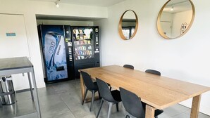 A conference room with a long wooden table, four chairs, a small table with a trash bin, and a vending machine.