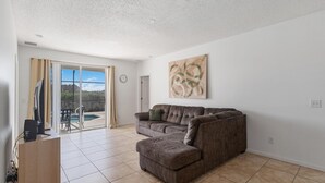 A living room with a large sectional sofa, a flat-screen TV on a wooden stand, and a sliding glass door leading to a balcony. - 5/3 Pool Home Located in Sunset Ridge Community! (Davenport)