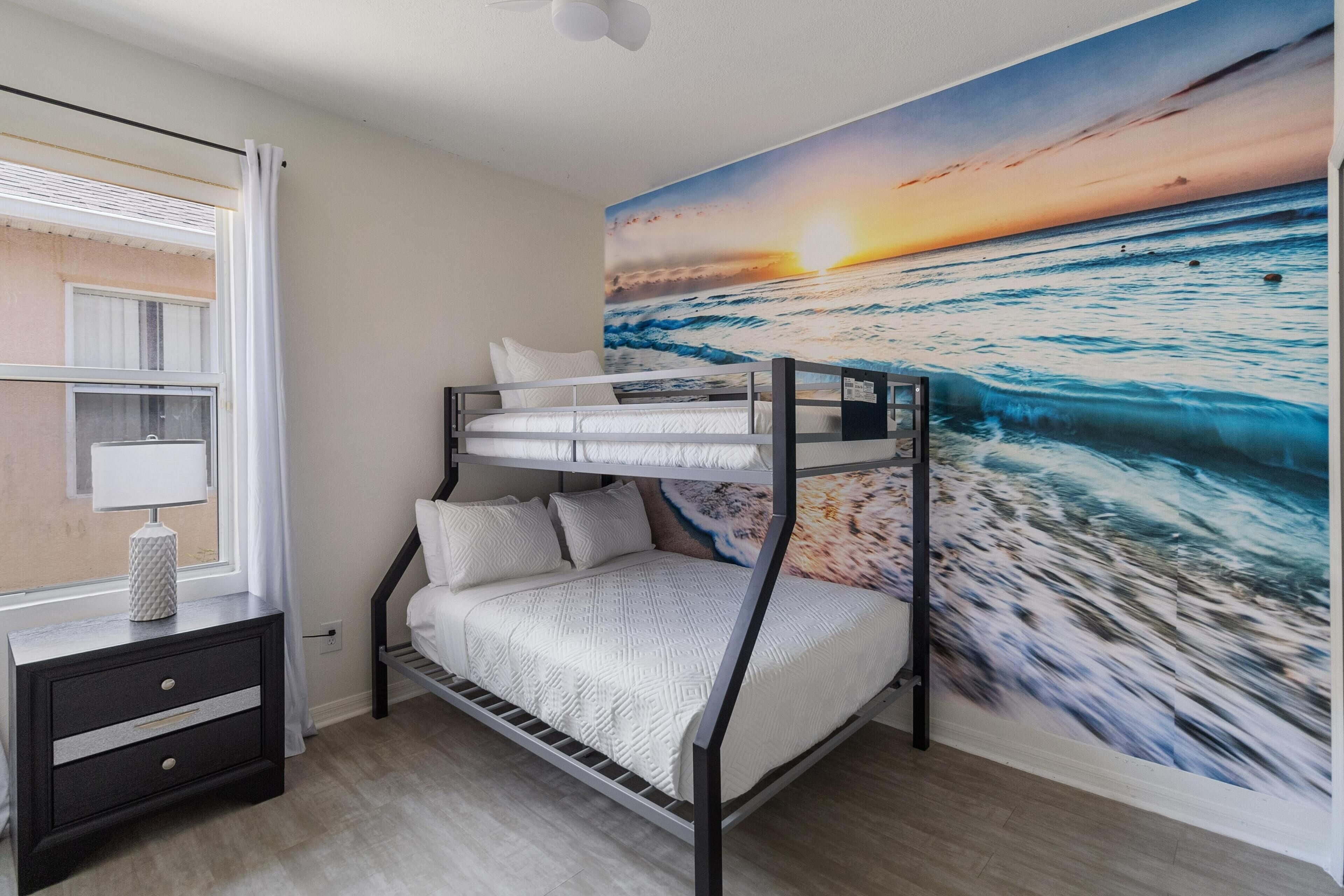 A bunk bed room with a sunset mural, a bedside table with a lamp, and a window with curtains.