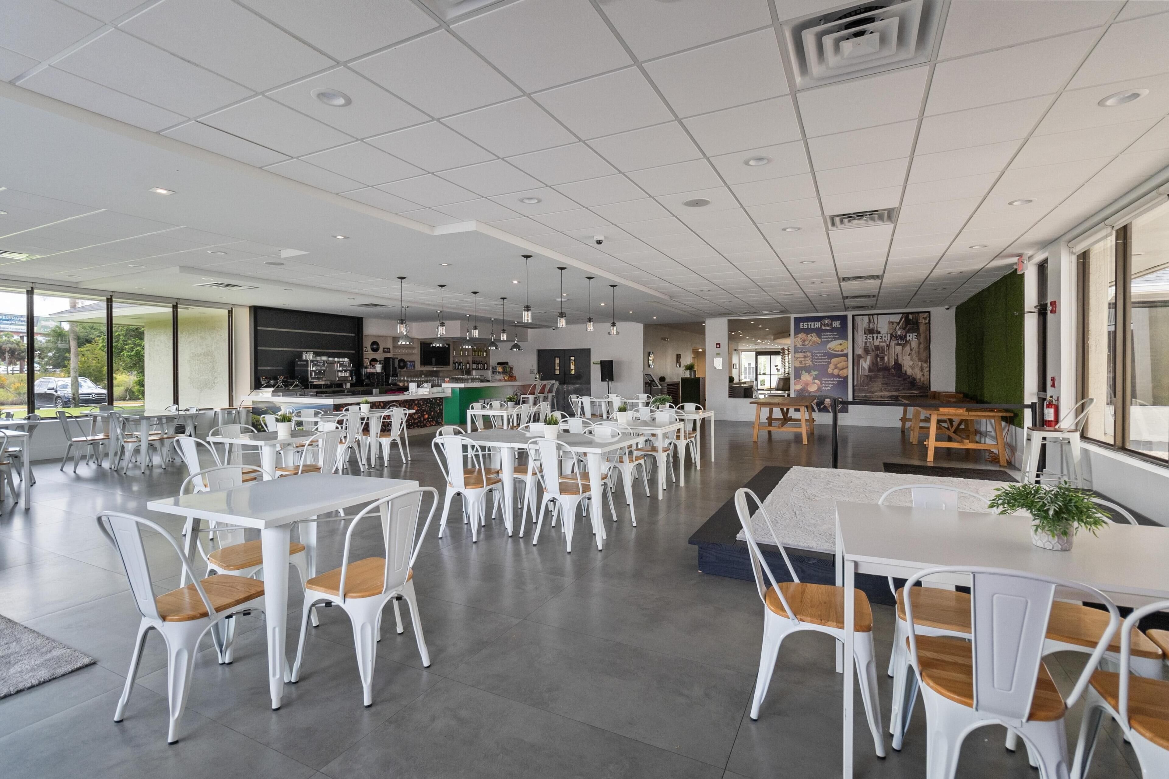 A spacious indoor dining area with white tables and chairs, a bar area, and large windows offering views of the outside.