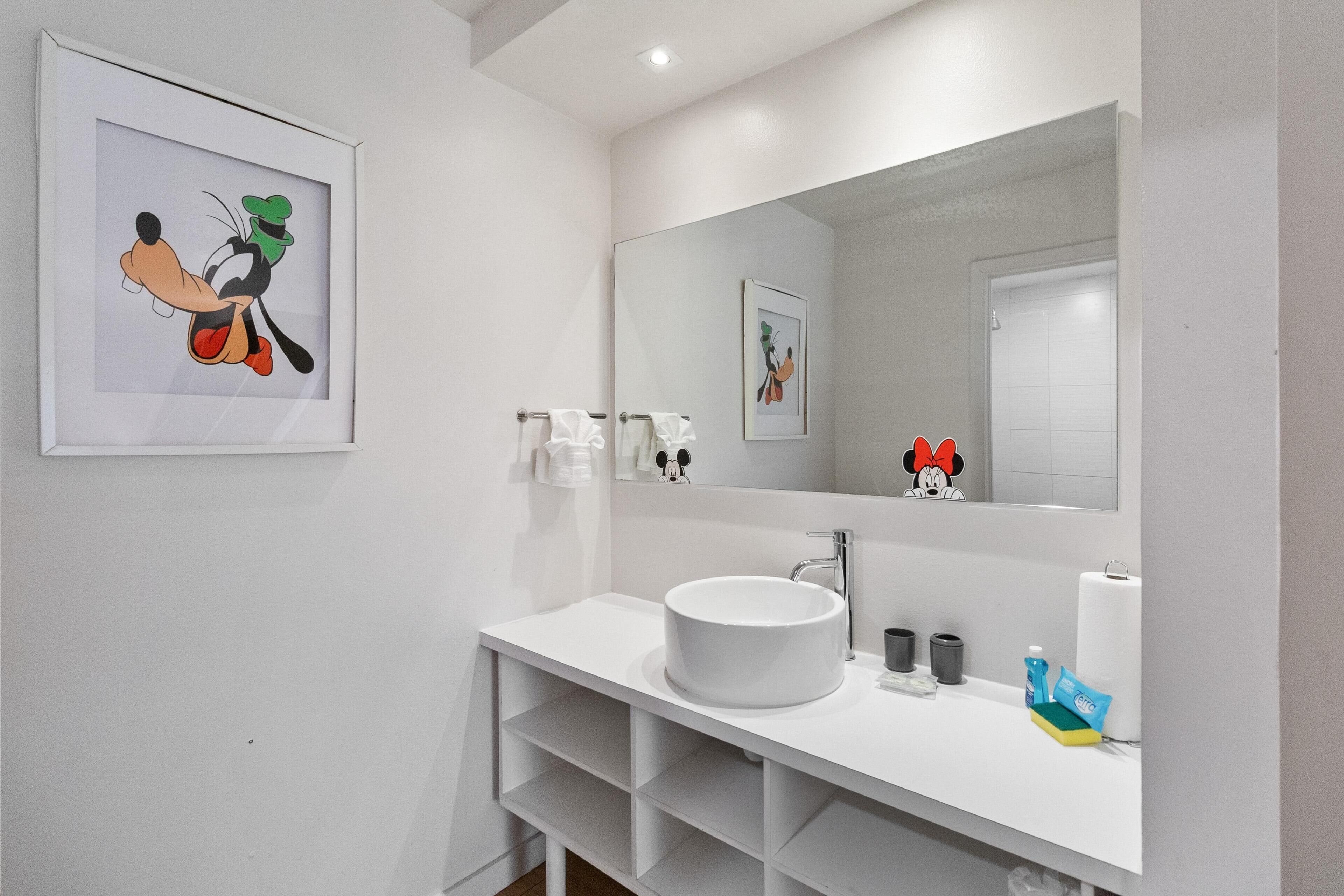 A modern bathroom with a white sink, mirror, and shelves. There are towels and toiletries on the counter.