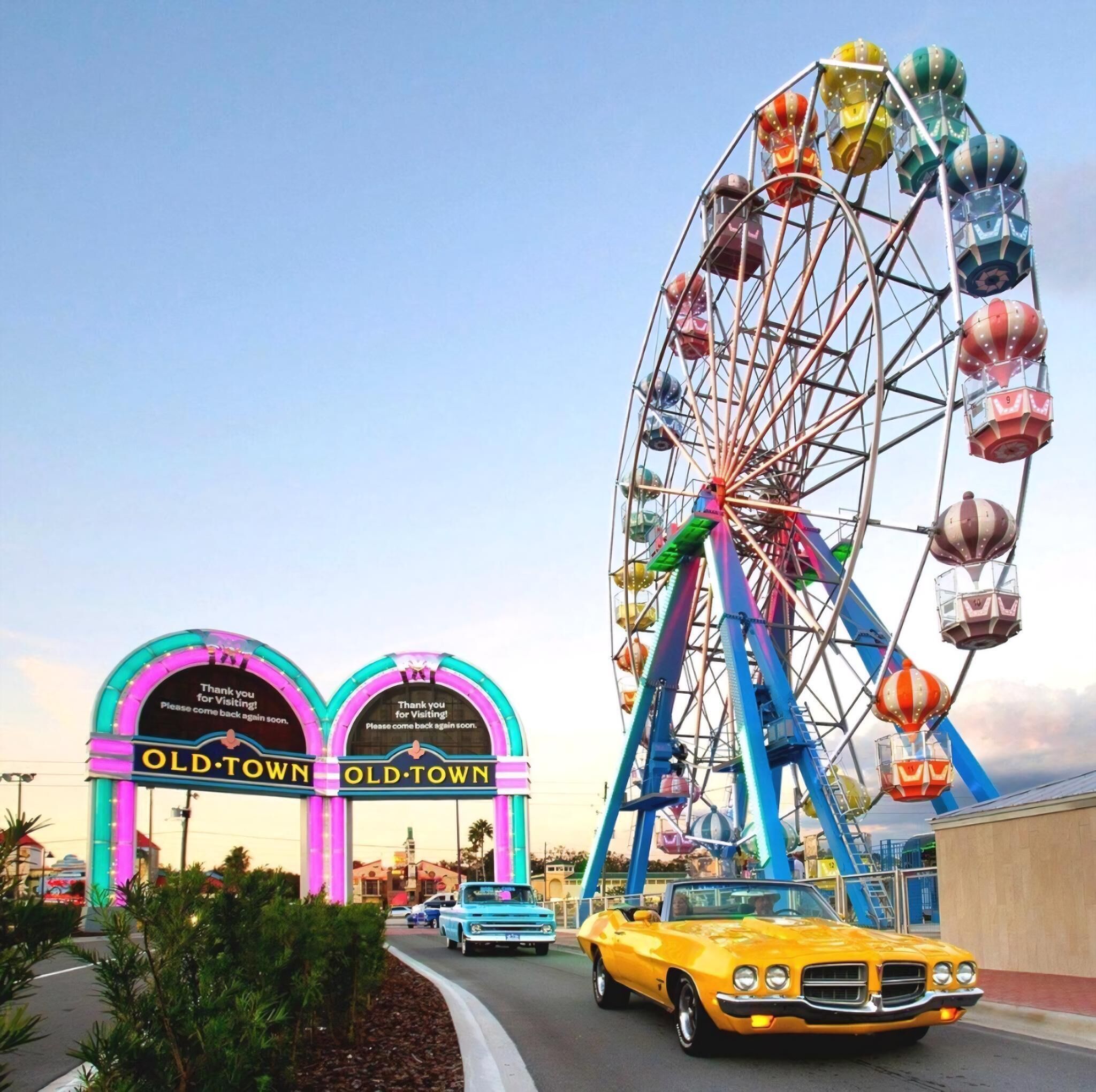 A Ferris wheel with colorful gondolas and a vintage car parked in front of a building with neon signs.