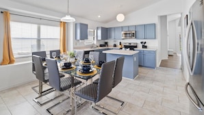 A modern kitchen with blue cabinets, a central island, and a dining area with a glass table and chairs. - Great Community, Amazing Private Pool, Near Disney! (Davenport)