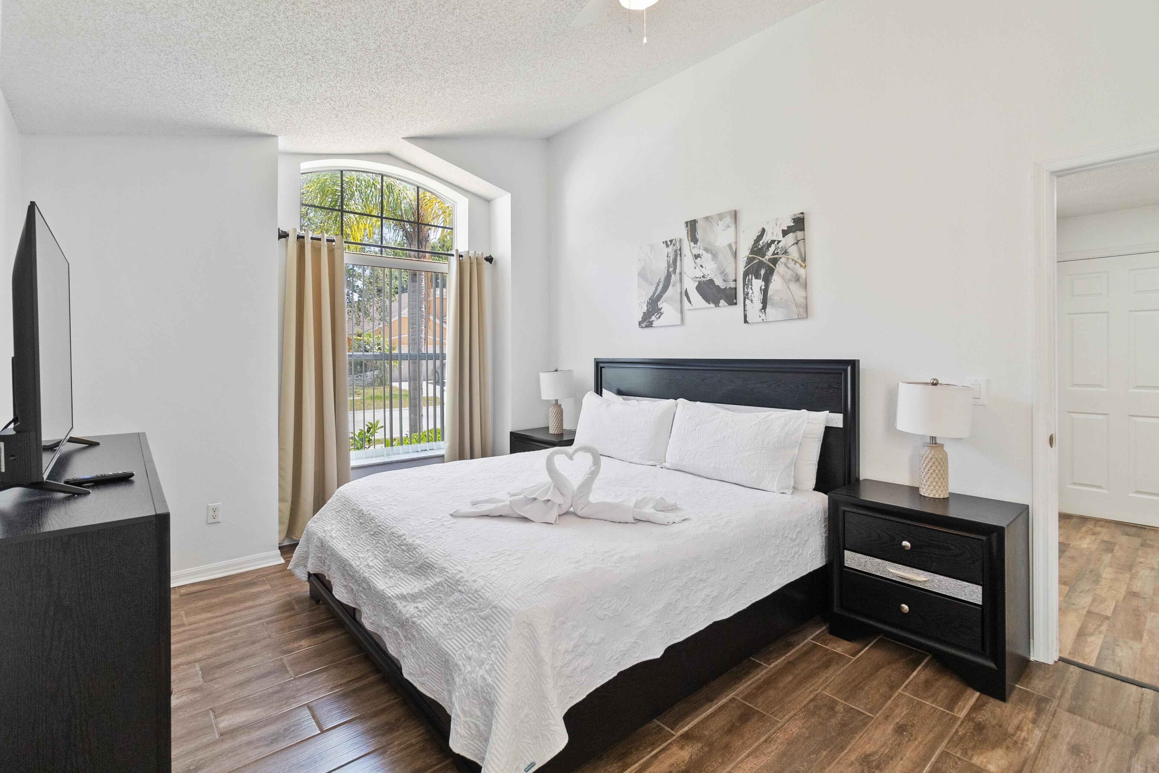 A modern bedroom with a large bed, a TV on a stand, and a window with curtains.