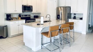 A modern kitchen with white cabinetry, stainless steel appliances, a granite countertop, and a central island with bar stools. - Amazing 4/3 Home at Reunion!! With Private Pool, Near Disney and Universal!! (Kissimmee)