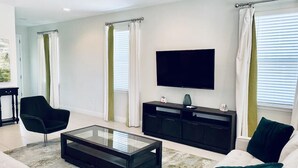 A modern living room with a glass coffee table, black entertainment unit, white sofa, and a flat-screen TV mounted on the wall. - Amazing 4/3 Home at Reunion!! With Private Pool, Near Disney and Universal!! (Kissimmee)