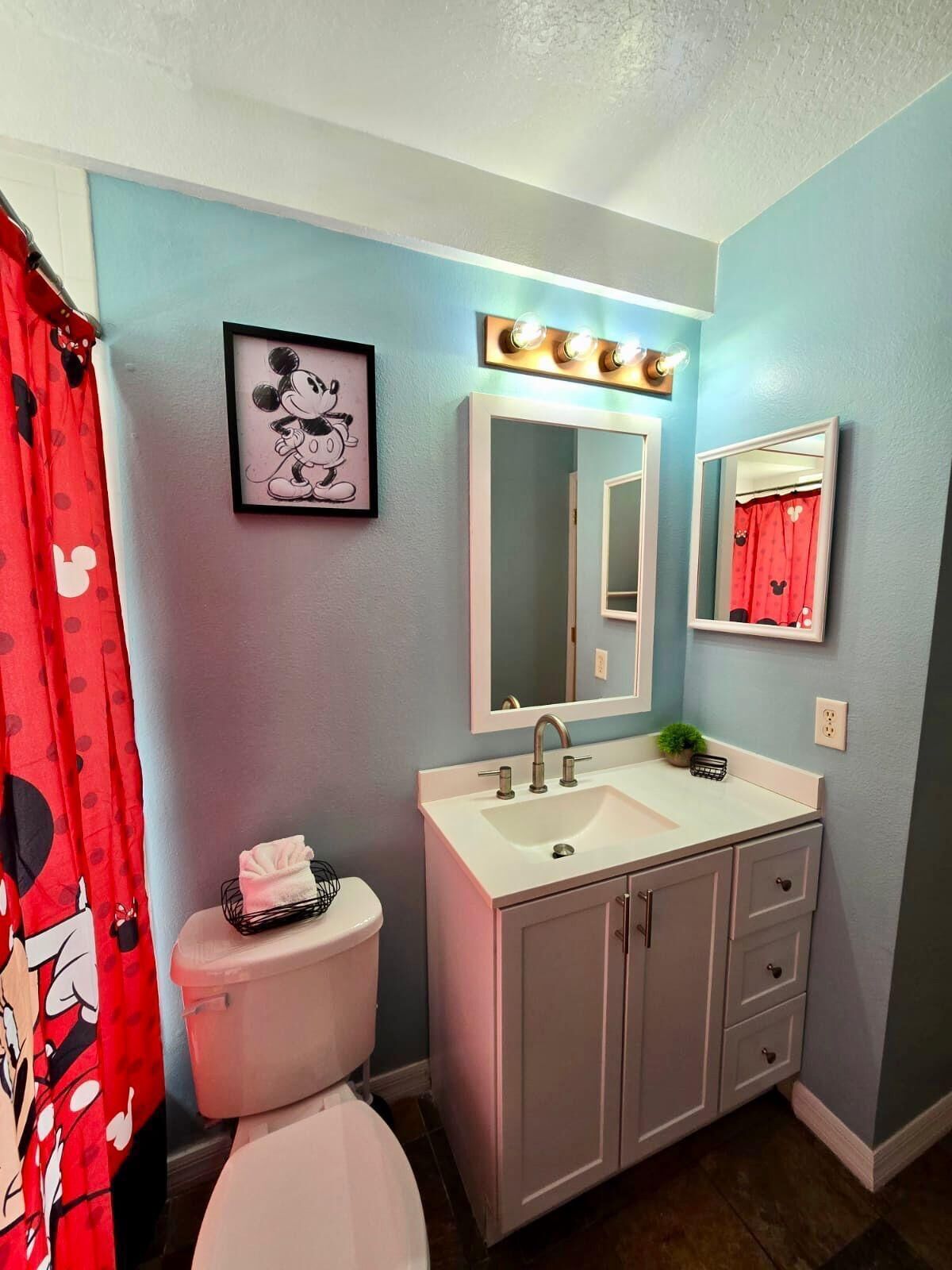 A bathroom with a white sink, a large mirror, and a Mickey Mouse framed picture on the wall.