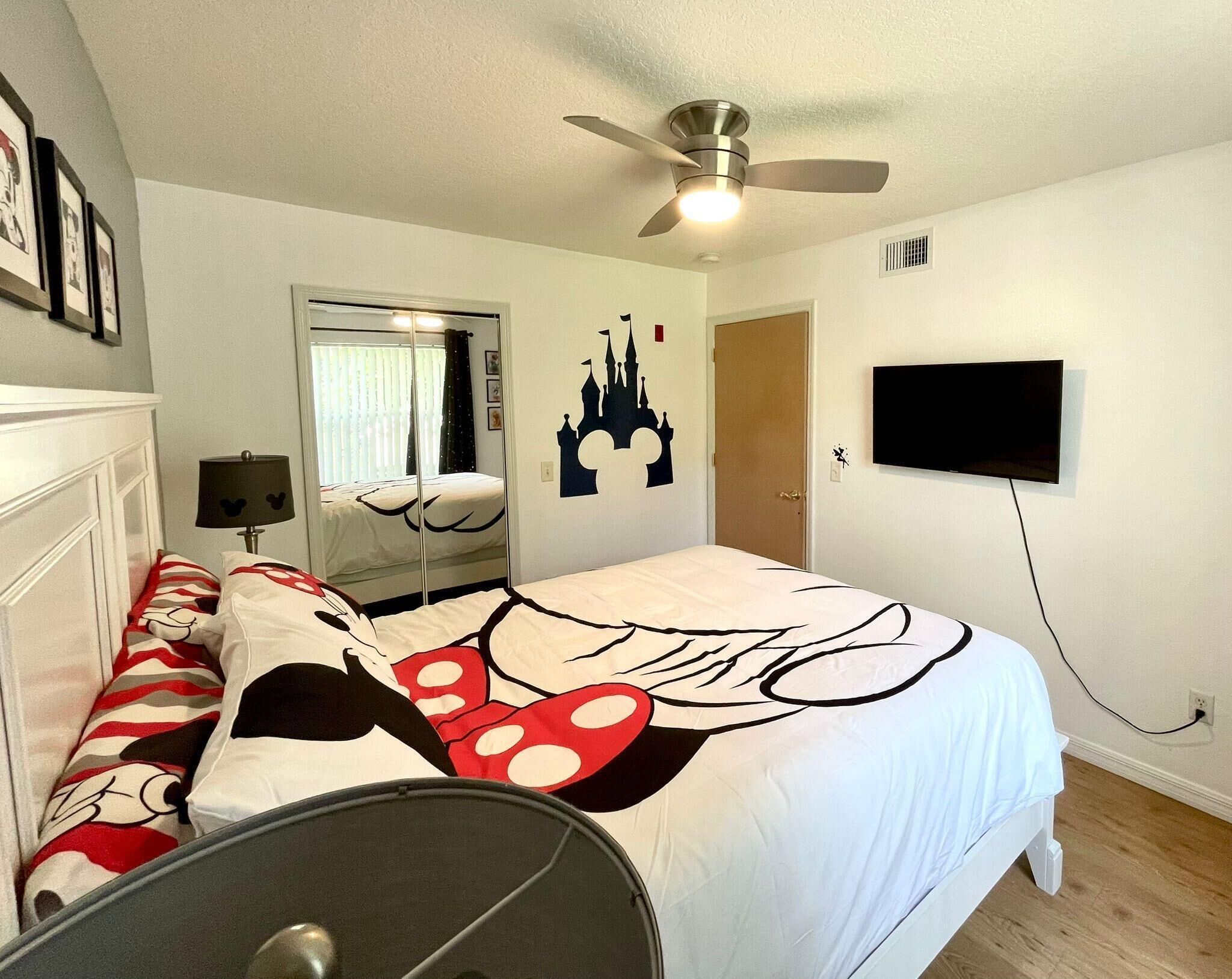 A bedroom with a bed, a ceiling fan, a TV mounted on the wall, and a window with curtains.