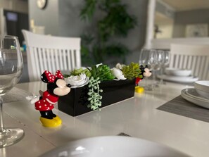 Et spisebord med en sort, aflang plantekasse med sukkulenter og hvide blomster, en Mickey Mouse-figur og et glas vand.