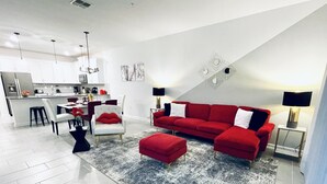 Een moderne woonkamer met een rode bank, een witte fauteuil en een eethoek, met een keuken op de achtergrond.