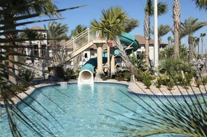 A swimming pool with a water slide, palm trees, and a building with a balcony in the background. - Brand New Condo at Champion Gate With Golf View, Near Famous Attractions (Kissimmee)