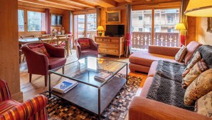A cozy wooden cabin interior with a dining area, a living room with a sofa and armchairs, a glass coffee table, and a television.