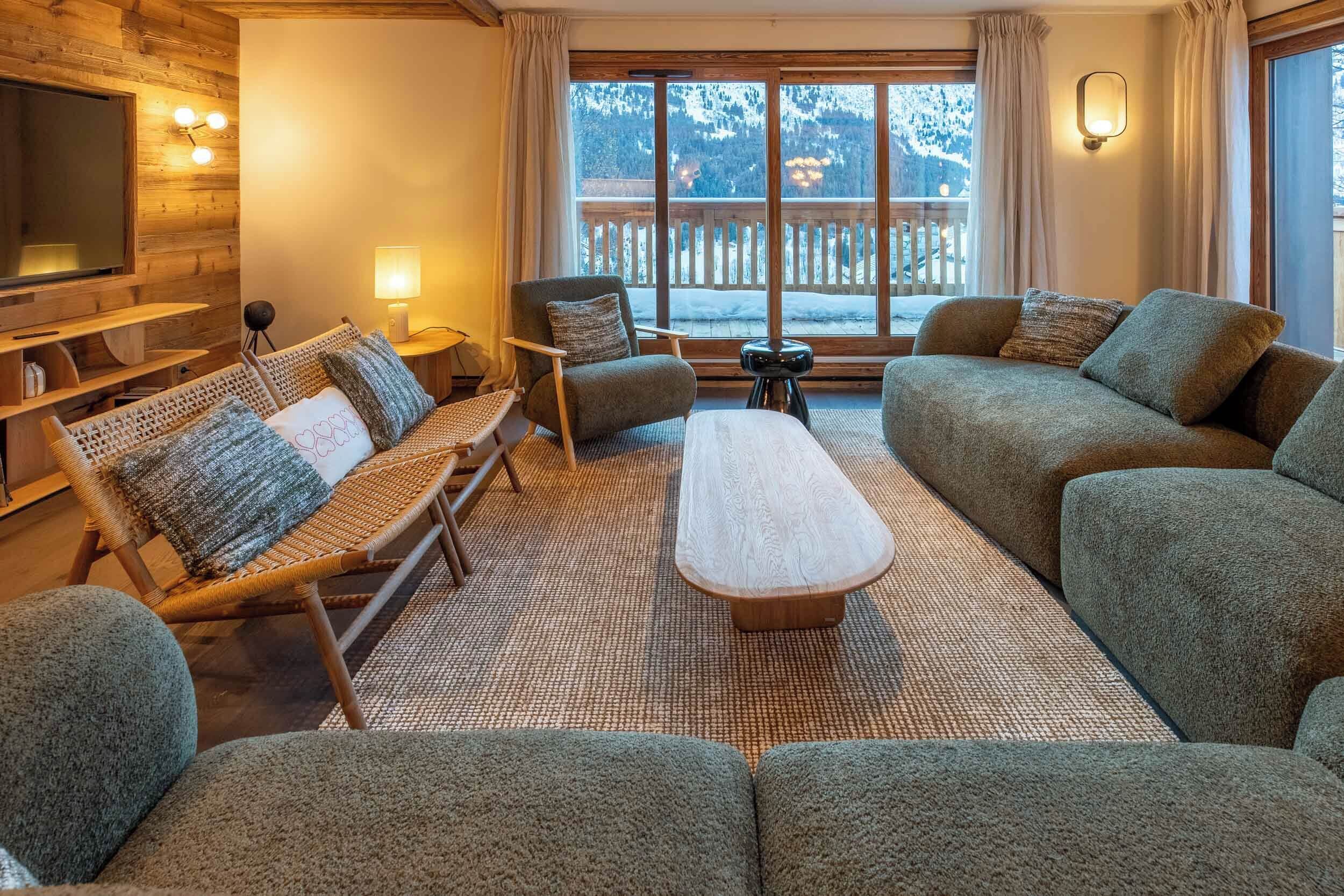A cozy living room with a sofa, armchair, and a coffee table. There is a TV mounted on the wall and a window with a view of the outdoors.