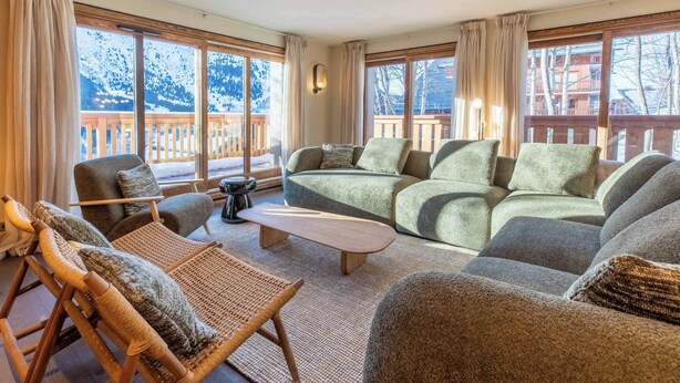 A cozy living room with a sofa, chairs, a small table, and a rocking chair. Large windows offer a view of a snowy mountain and a wooden deck outside.