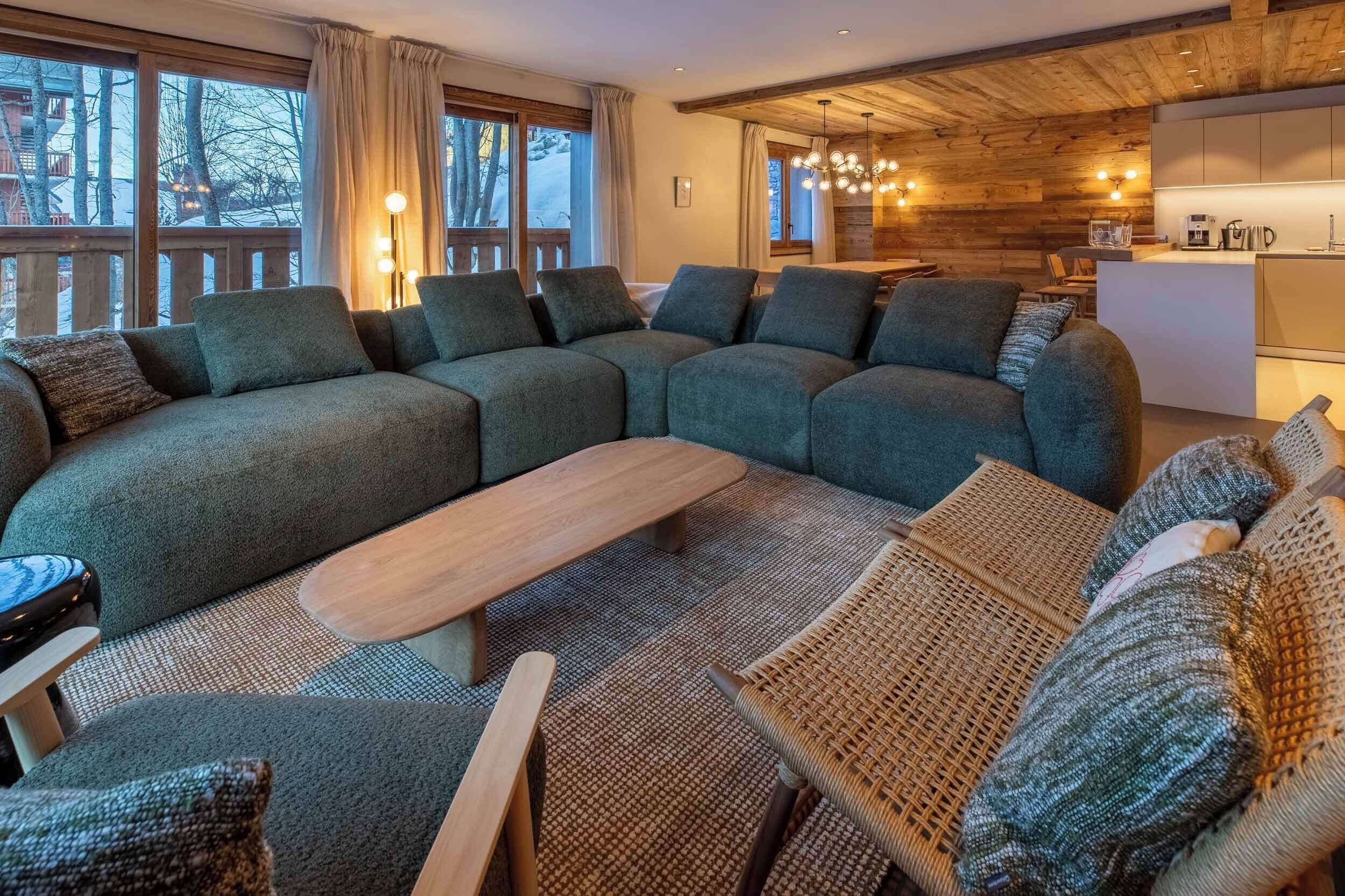 Una sala moderna con un sofá seccional grande, una mesa de centro de madera y sillas de mimbre. Se ve un área de cocina al fondo.
