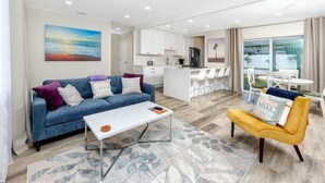A modern living room with a blue sofa, a yellow armchair, and a white coffee table. There is a kitchen area with white cabinets and a dining area with white chairs.