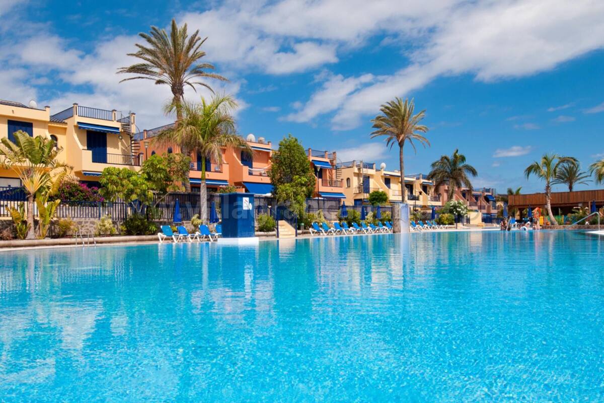A large swimming pool with lounge chairs and palm trees, surrounded by multi-story buildings with balconies.