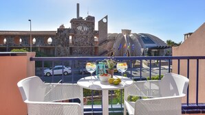 A balcony with two white wicker chairs, a small round table with a bowl of fruit and two glasses of water, and a view of a building with a clock tower and arches. - Meloneras Duplex (San Bartolomé de Tirajana)