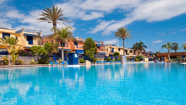 A clear blue pool with lounge chairs and palm trees, surrounded by buildings with balconies and a clear sky. - Meloneras Duplex (San Bartolomé de Tirajana)