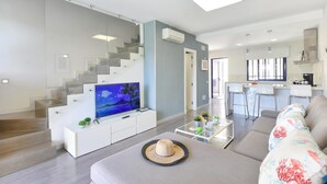 A modern living room with a flat-screen TV, a sofa, a coffee table, and a staircase leading to a kitchen area. - Meloneras Duplex (San Bartolomé de Tirajana)