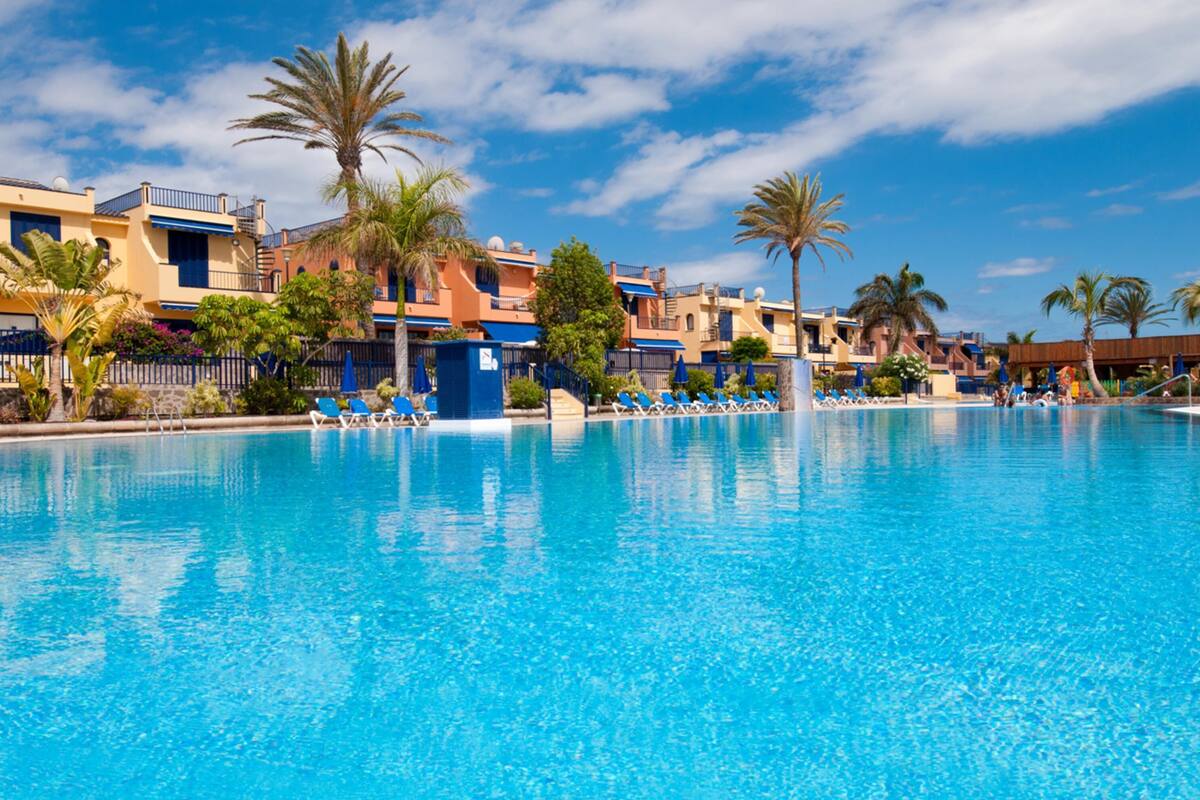 A swimming pool with lounge chairs and palm trees, surrounded by multi-story buildings with balconies.