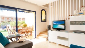 A modern living room with a TV, white furniture, a balcony with outdoor seating, and a view of a building with arches. - Meloneras Duplex (San Bartolomé de Tirajana)