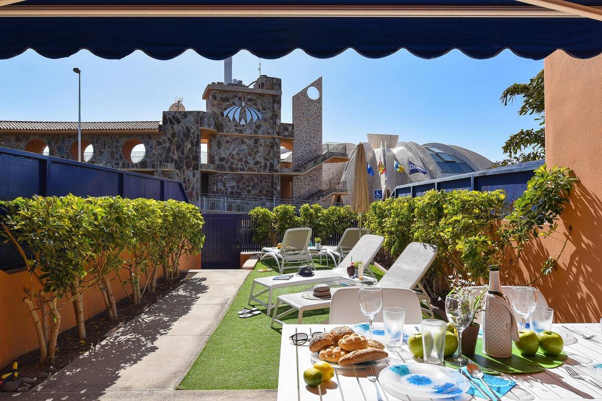An outdoor dining area with a table set for two, surrounded by lounge chairs and a decorative umbrella.