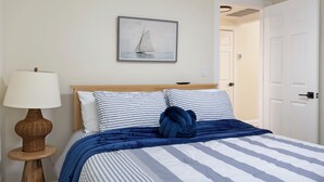 Un letto rifatto con lenzuola a righe e una coperta blu, un quadro appeso al muro raffigurante una barca a vela e un comodino in legno con una lampada.