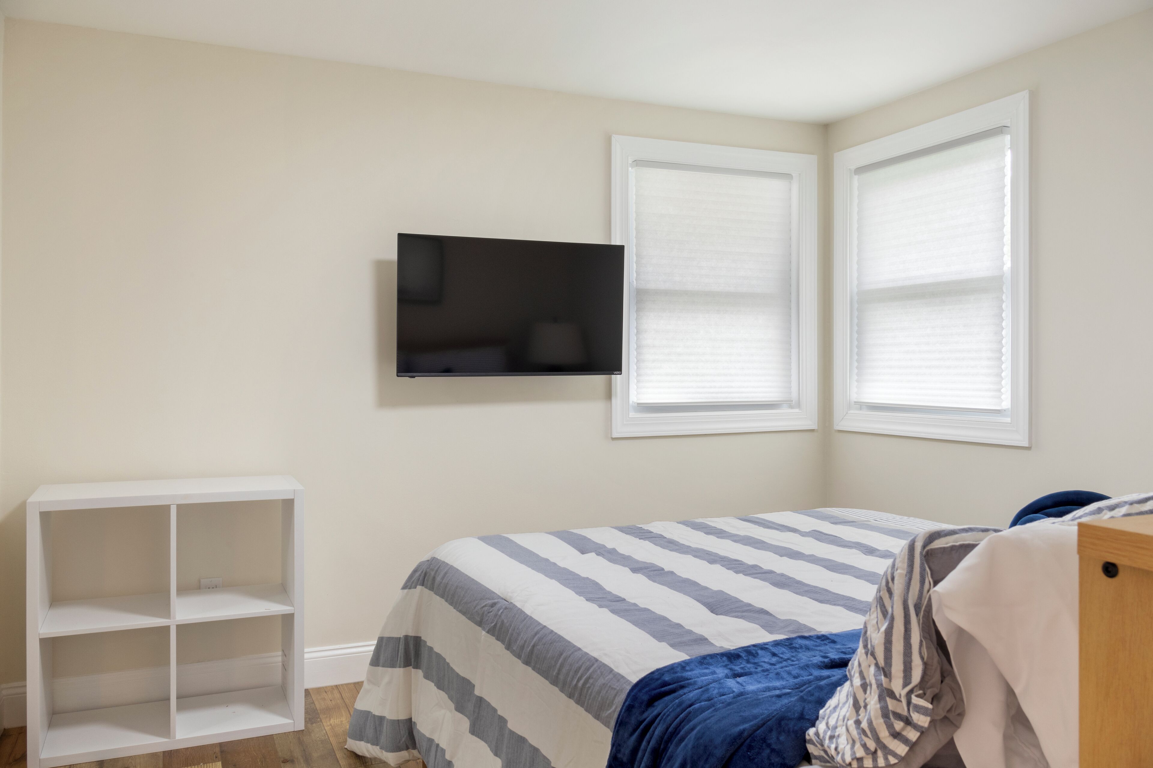 Un dormitorio con cama, televisor montado en la pared y dos ventanas con persianas.