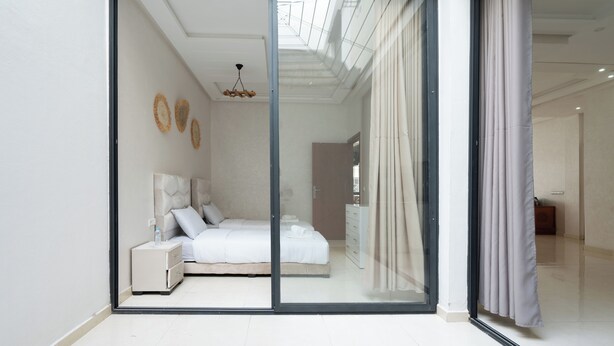 A modern bedroom with a glass door, a bed with white linens, a nightstand, and a ceiling light. - Apartment Near the Beach (Bouznika)