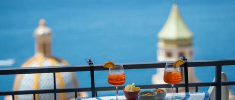 A table with two drinks and snacks, overlooking a coastal view with a distinctive building in the background.