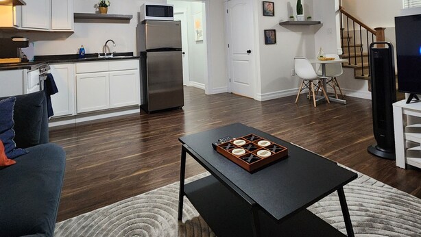 A modern living room with a dark wood floor, a black coffee table, and a blue sofa. A kitchen with white cabinets and stainless steel appliances is visible in the background. - Private apartment in Moncton (Moncton)