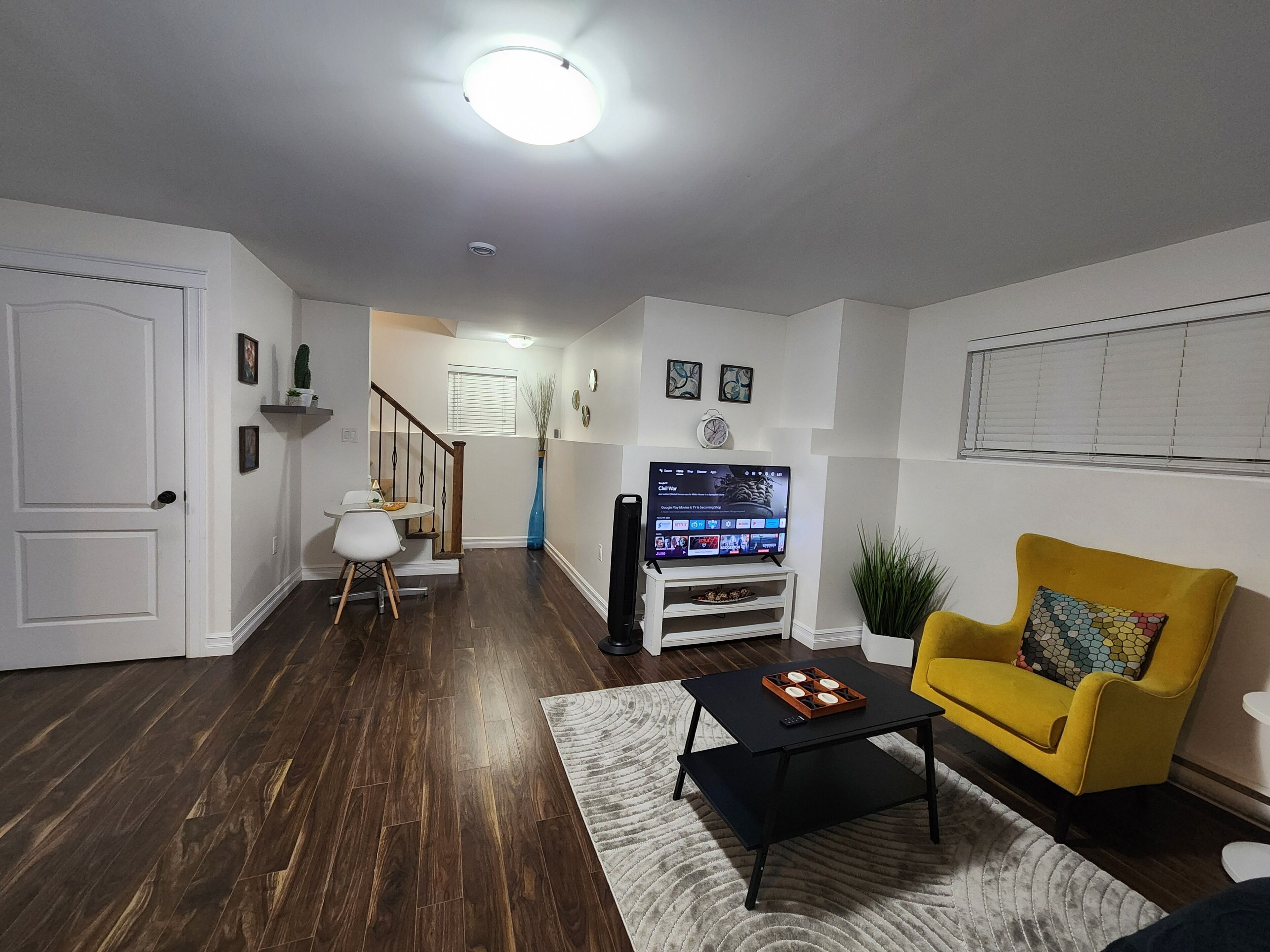 A living room with a wooden floor, a yellow armchair, a black coffee table, a TV, and a staircase leading to another room.