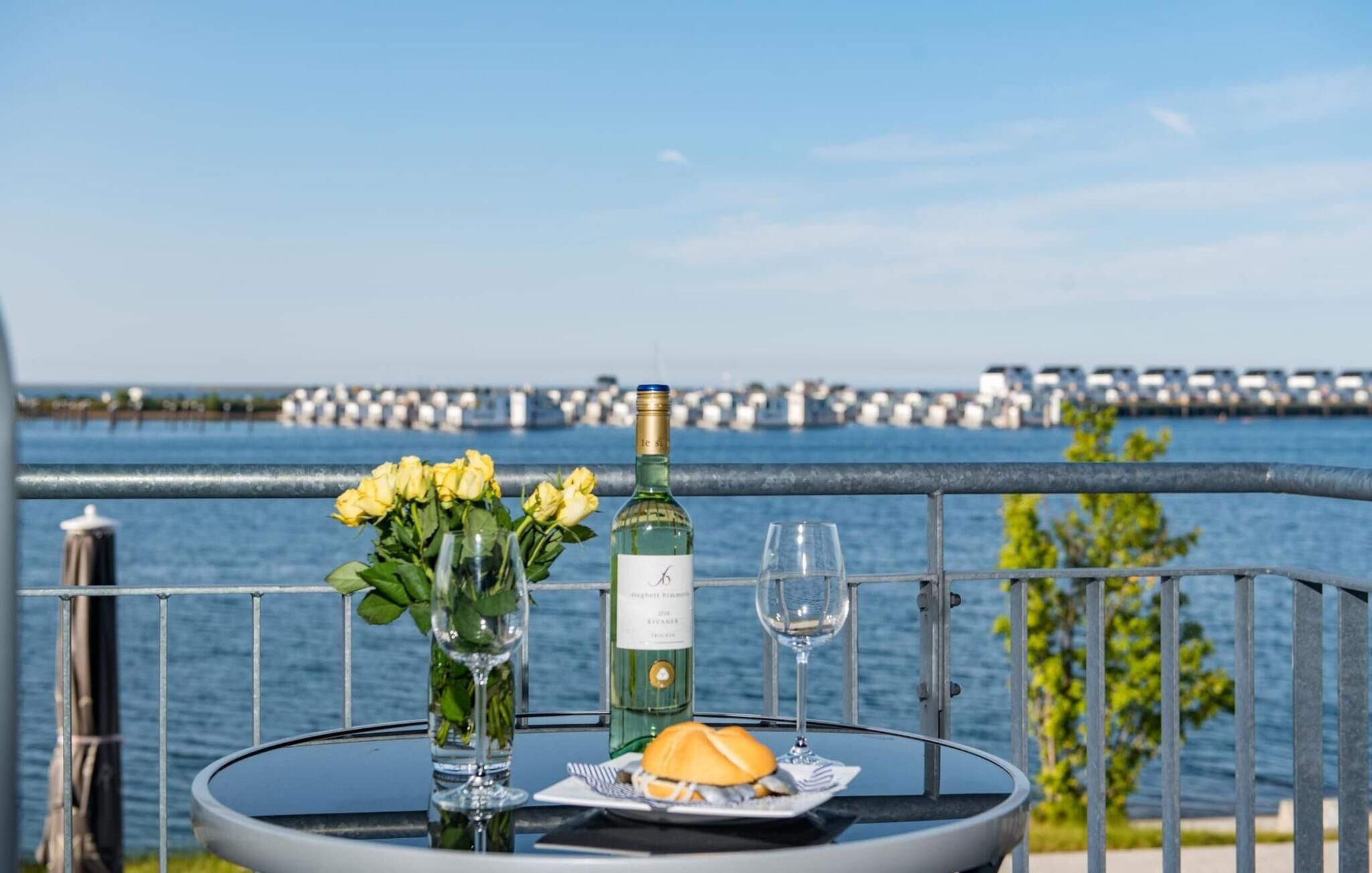 A table with a bottle of wine, a glass, and a plate with food, set against a scenic waterfront view.