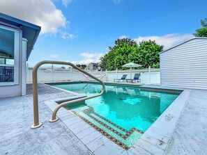 Ein Swimmingpool mit gefliestem Deck, einer Metallleiter und Liegestühlen unter einem Baum.