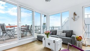 Een ruime woonkamer met grote ramen, een grijze bank, een witte opbergkast en een paarse fauteuil.