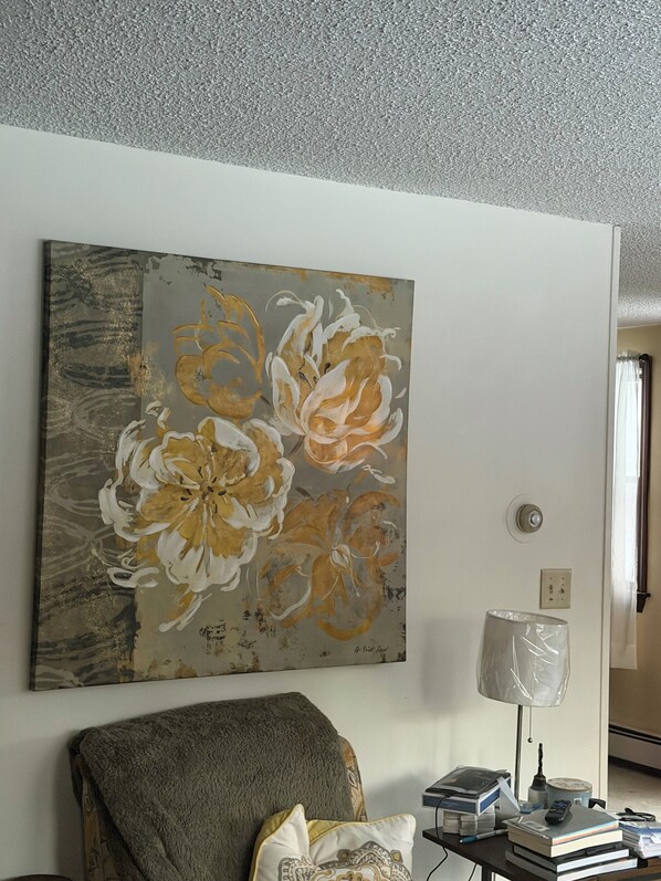 Una sala de estar con un cuadro grande de flores en la pared, un sillón gris, una lámpara y una mesa con libros y un control remoto.