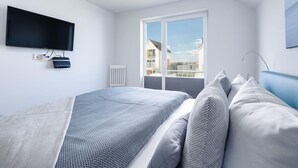 A modern bedroom with a large bed, a flat-screen TV mounted on the wall, and a window offering a view of buildings. - Gorgeous apartment in Olpenitz (Olpenitz)