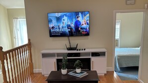 A living room with a flat-screen TV mounted on the wall, a dark wooden coffee table with two potted plants, and a grey sofa. - Charming 3-bedroom house. (Cortlandt)