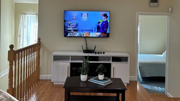 A living room with a flat-screen TV mounted on the wall, a wooden staircase, a white entertainment unit, and a bed in the adjacent room. - Charming 3-bedroom house. (Cortlandt)