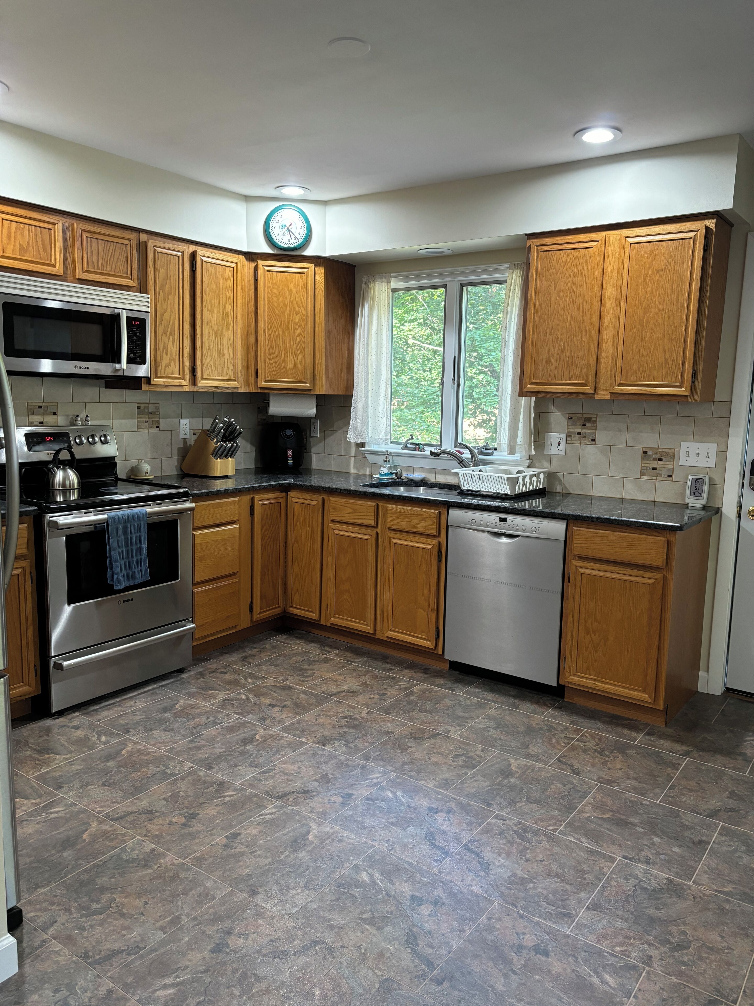 A kitchen with wooden cabinets, a Bosch microwave, a stainless steel oven, a dishwasher, and a window with a view of greenery.