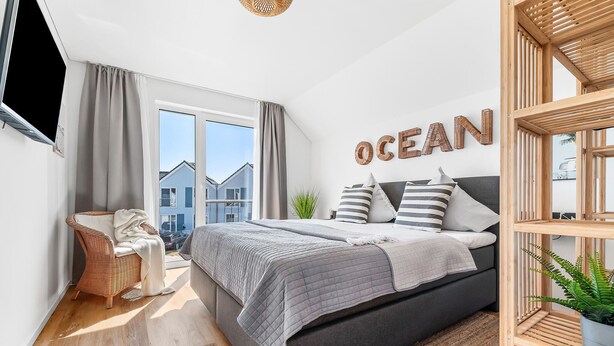 Une chambre moderne avec un grand lit, une literie grise et une vue sur les maisons voisines, visible à travers une grande fenêtre.