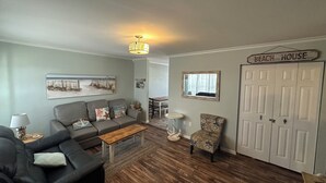 A living room with a grey sofa, a wooden coffee table, a patterned armchair, and a 'BEACH HOUSE' sign on the wall.