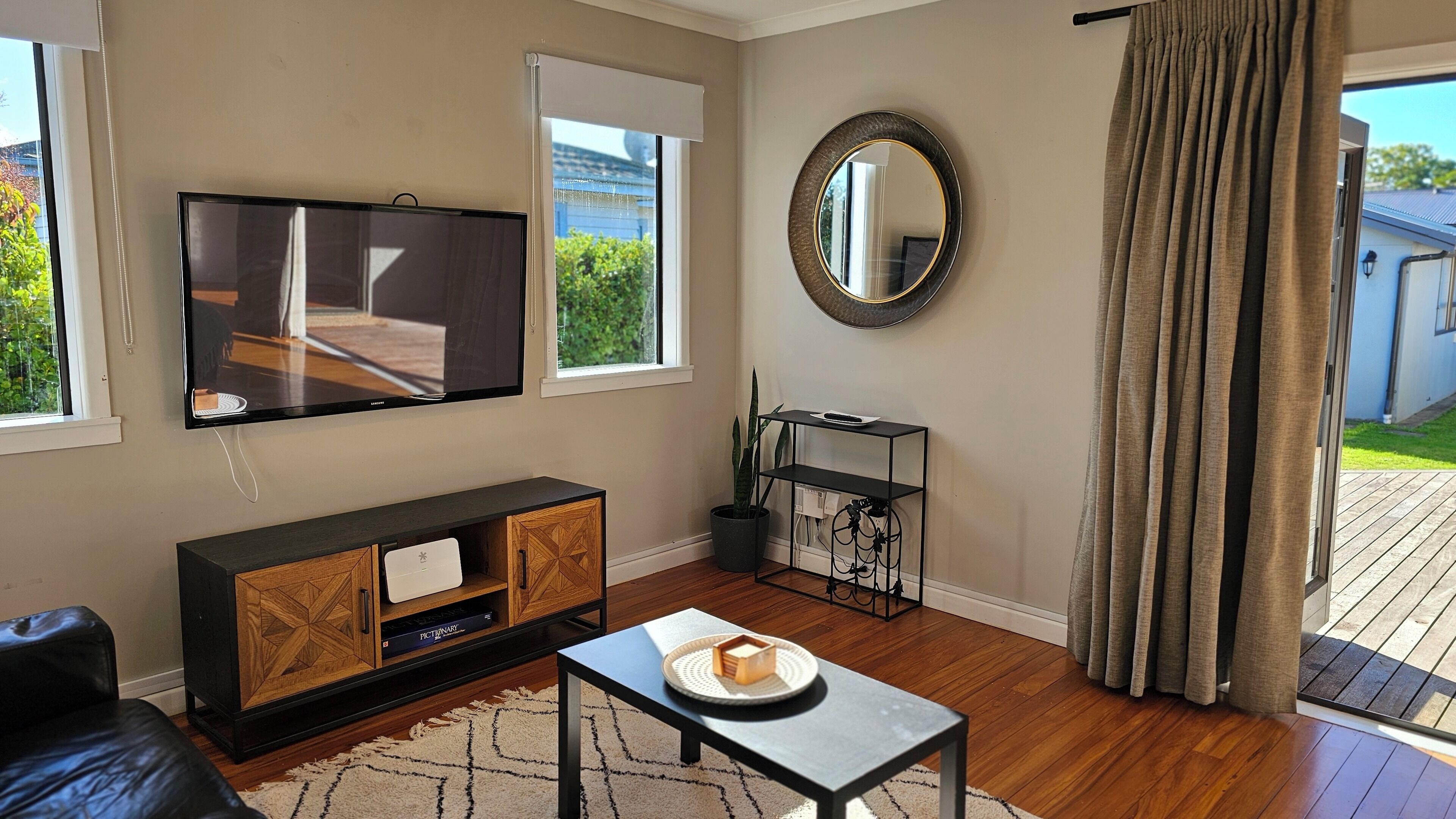 A living room with a flat-screen TV, a round mirror, a wooden cabinet, a small table with a plate, and a view of a house through the window.