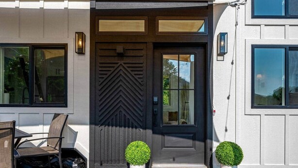Ein modernes Haus mit einer dunklen Haustür, zwei Blumentöpfen, einer Bank und einer Fußmatte mit dem Aufdruck 'hello'.