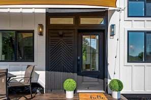 A modern house with a dark front door, two potted plants, a bench, and a 'hello' mat.